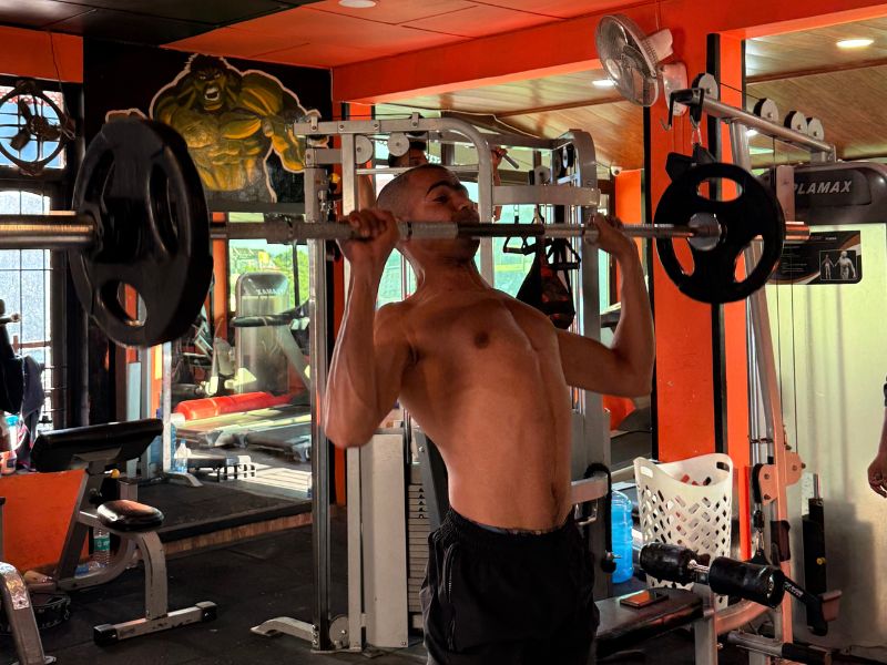 Man performing standing barbell shoulder press in Dharamkot Man performing standing barbell shoulder press in Dharamkot, focusing on strength training with weight plates on both sides of the bar